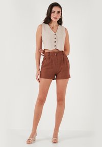 Gilet en lin beige coupé avec des boutons, associé à un short marron taille haute. Modèle portant des sandales à lanières, cheveux coiffés en vagues.
