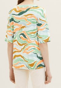 Femme portant un haut ample à manches courtes avec des rayures ondulées abstraites en vert, orange, moutarde et bleu, associé à un pantalon de couleur claire.