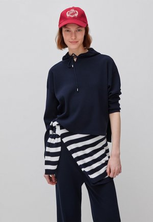 Junge Frau mit roter Kappe, marineblauem Hoodie und marineblauen Hosen, die einen marineblau-weiß gestreiften Pullover um die Taille gebunden hält.