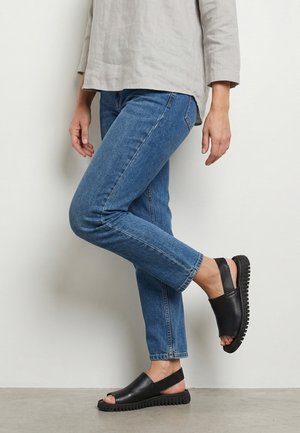 Persoon die een blauwe spijkerbroek, een beige top met lange mouwen en zwarte open-teen slip-on sandalen met dikke zolen draagt, staat op één been tegen een witte muur.