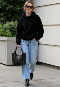 Felpa nera con cappuccio, jeans larghi azzurri, borsa a tracolla in pelle nera e stivali neri con tacco a punta, su uno sfondo neutro.