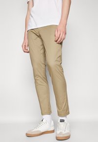 Chino beige clair avec une coupe slim, présentant une texture lisse et un design simple. Associé à des baskets blanches et des chaussettes blanches jusqu'aux chevilles.