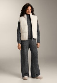 Frau trägt eine cremefarbene Fleeceweste über einem dunkelgrauen Strickrollkragenpullover und weiten Hosen mit beigefarbenen Plateau-Schuhen, stehend vor einem neutralen Hintergrund.