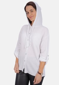 Weißes, strukturiertes Kapuzenshirt mit Knopfleiste, hochgekrempelten langen Ärmeln und Kordelzug am Hals, kombiniert mit schwarzen Kunstlederhosen.