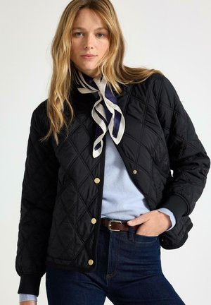 Kvinde iført en sort quiltet jakke, lyseblå sweater, mørke jeans, brunt bælte og sort-hvidt stribet halstørklæde.