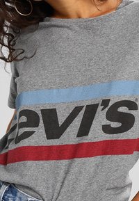 Camiseta de algodón gris con el logo de "Levi's" en negrita negra, acentuada por franjas horizontales azules y rojas a lo largo del pecho. Mangas cortas.