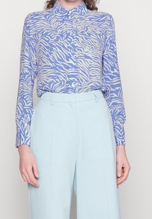 Femme portant une chemise à manches longues boutonnée avec un imprimé animal bleu et blanc, rentrée dans un pantalon taille haute bleu clair.
