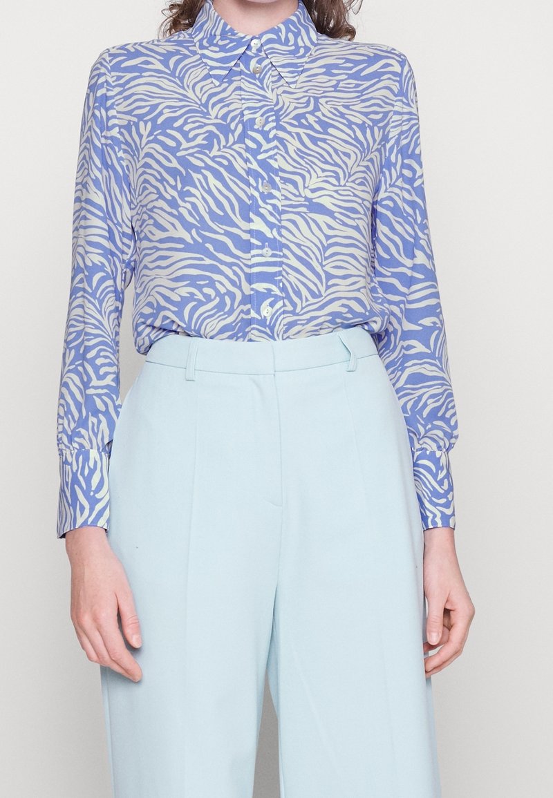 Femme portant une chemise à manches longues boutonnée avec un imprimé animal bleu et blanc, rentrée dans un pantalon taille haute bleu clair.
