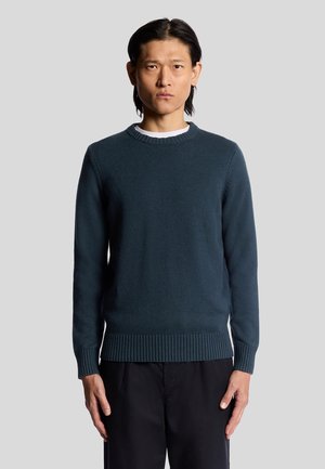 Dark teal knitted sweater with crew neckline, ribbed cuffs and hem, featuring a smooth texture and a classic fit. Worn over a white shirt.