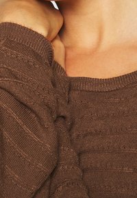 La mano di una persona riposa vicino al collo, indossando un maglione di maglia marrone con motivi orizzontali testurizzati e un dettaglio a coste sul collo.