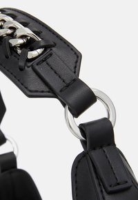 Sort læderrem med sølvfarvet hardware, stikninger og en cirkulær ringfastgørelse. Tekstureret overflade og fremtrædende snoredesign.