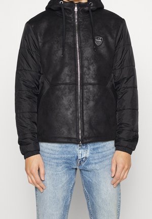 Homme portant une veste noire à capuche avec des manches matelassées et le logo EA7, associée à un jean bleu clair sur un fond uni.