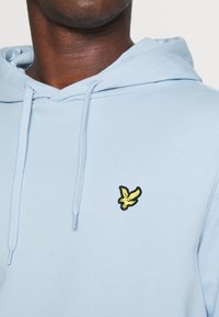 Sudadera con capucha de color azul claro, hecha de algodón, con un logo de pájaro bordado en negro y amarillo en el lado izquierdo del pecho. Textura suave, ajuste casual.