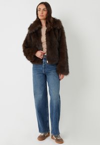 Braune Kunstfelljacke mit breitem Kragen, langen Ärmeln und taillierter Passform, kombiniert mit blauen geraden Jeans und braunen Sneakern.