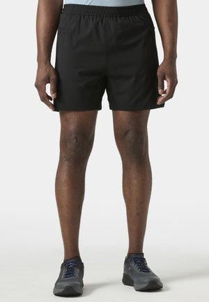 Man in zwarte sportshorts en grijze hardloopschoenen, staand tegen een effen lichte achtergrond.