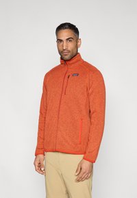 Ζακέτα από φλις σε ζωντανό πορτοκαλί χρώμα με ψηλό γιακά, πλήρες φερμουάρ και κόκκινες λεπτομέρειες. Διαθέτει πλαϊνές τσέπες και μικρό λογότυπο στο στήθος.