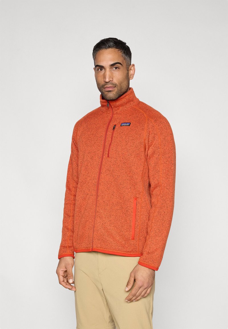 Ζακέτα από φλις σε ζωντανό πορτοκαλί χρώμα με ψηλό γιακά, πλήρες φερμουάρ και κόκκινες λεπτομέρειες. Διαθέτει πλαϊνές τσέπες και μικρό λογότυπο στο στήθος.