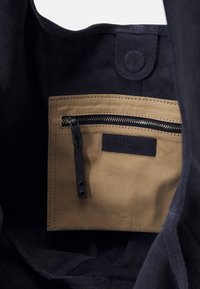 Vue intérieure d'un sac montrant une poche zippée en tissu beige avec un tirette en cuir noir et une fermeture à bouton-pression au-dessus.