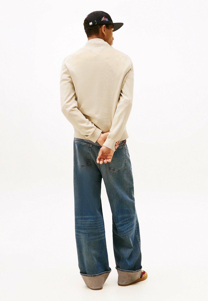 Suéter de punto beige con un cuello bajo, combinado con jeans azules holgados. Presenta una textura lisa y un ligero desteñido. Zapatos grises con una suela marrón.