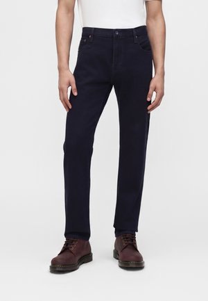 Homme portant un jean bleu marine foncé, une chemise blanche et des bottes à lacets marron foncé, debout devant un fond clair uni.