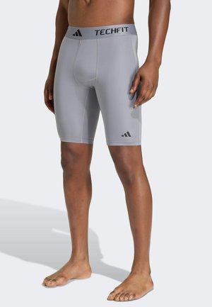 Homme portant un short de compression gris Adidas Techfit, debout pieds nus, une main détendue le long du corps.