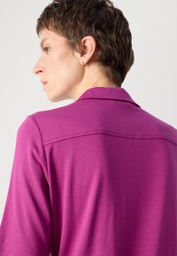 Fuksia polo skjorte med myk tekstur, korte ermer og krage; har en sydd sømdetalj over ryggen.