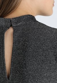 En kvinnas nacke och axel som bär en glittrig svart topp med hög hals och en nyckelhålsöppning bak som är snabbad med en liten knapp.