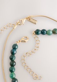 Goldfarbenes Halskettchen mit runden, facettierten grünen Perlen und einem Karabinerverschluss. Die Kette verleiht verstellbare Länge und einen dezenten Glanz.