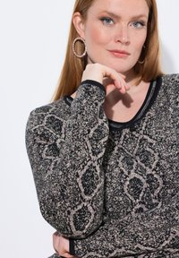 Sweter dzianinowy z czarno-beżowym wzorem geometrycznym, okrągłym dekoltem i czarnym wykończeniem. Model ma na sobie dużą biżuterię nauszną.
