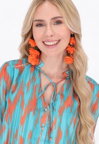 Turquoise és narancssárga mintás blúz, elöl megkötő résszel, V-alakú nyakkivágással és gombos díszítéssel. Élénk narancssárga virágos fülbevalók kiemelkedő részletként szolgálnak.