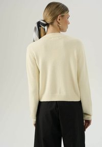 Maglione di colore crema, con texture, maniche lunghe e colletto a coste, abbinato a pantaloni neri. Capelli legati con un accessorio a fascia bianco e nero.