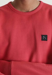 Sudadera roja con un cuello redondeado acanalado y puños. Presenta un pequeño parche cuadrado negro con un diseño gráfico blanco. Textura de tela suave.