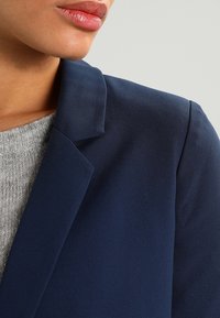 Blazer azul marino con solapa de muesca, hecho de una tela suave. Debajo se ve un suéter de punto gris, que complementa el color del blazer.