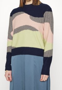 Femme portant un pull à motif abstrait multicolore avec des sections bleu marine, rose, gris et vert clair, associé à une jupe bleue.
