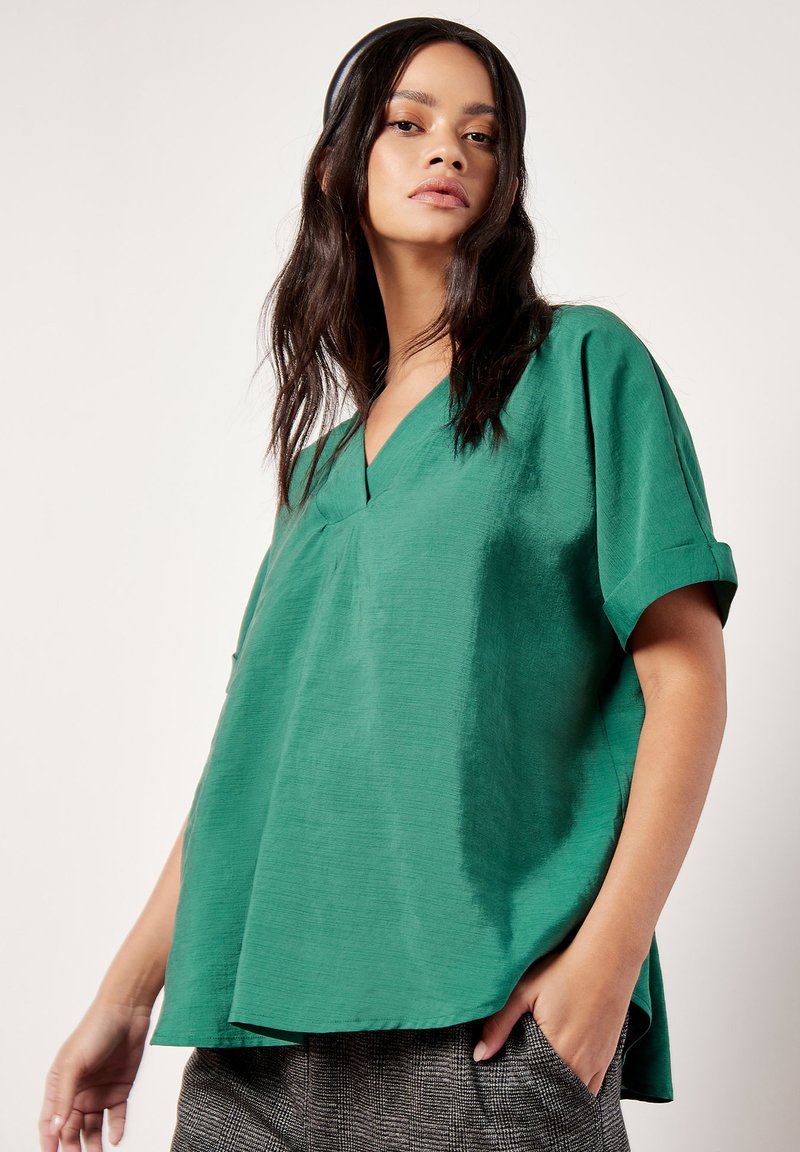 Top verde a maniche corte con scollatura a V, realizzato in un tessuto testurizzato. Vestibilità ampia con orlo curvo. Abbinato a pantaloni grigi con fantasia.