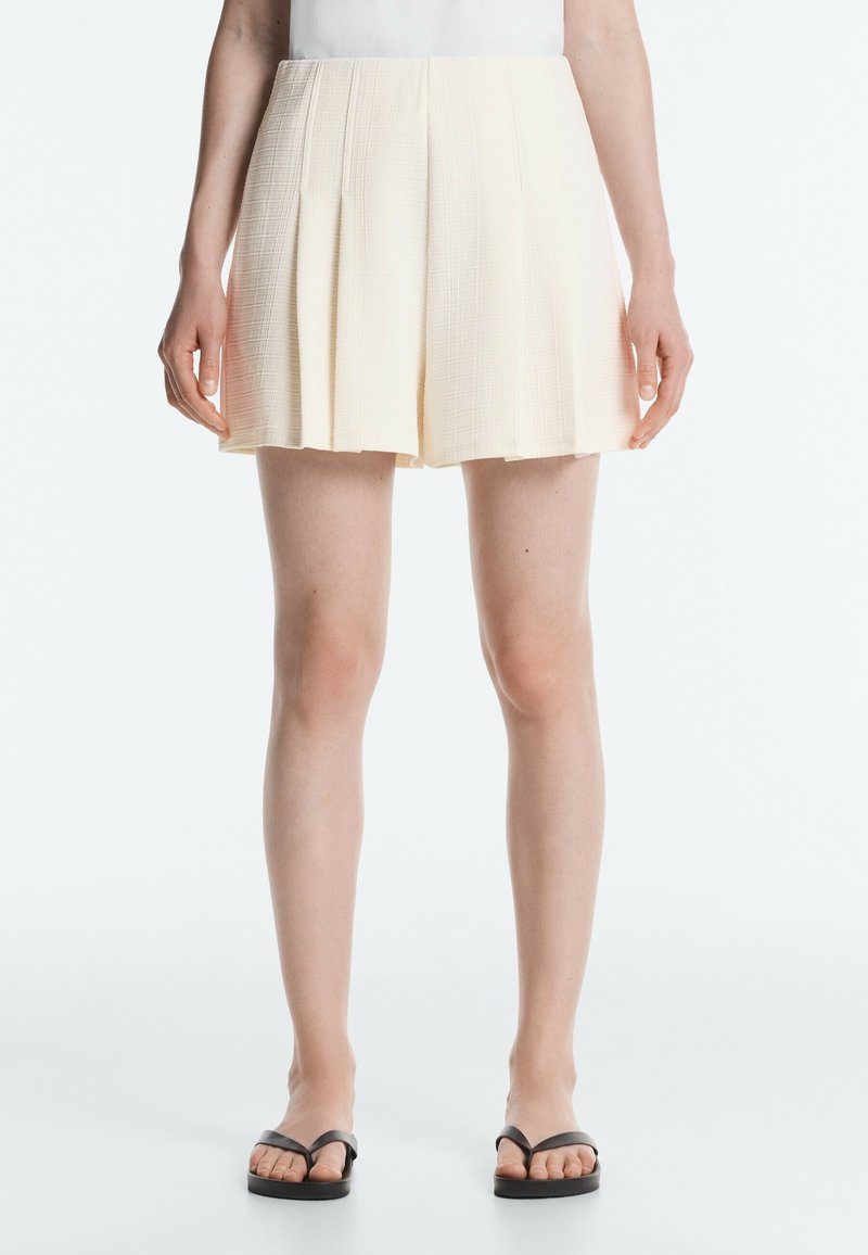 Femme portant un short plissé crème, un haut blanc et des sandales noires, debout devant un fond clair uni.