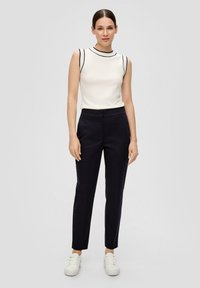 Mouwloos wit topje met zwarte rand, gecombineerd met slim-fit donkerblauwe broek en witte sneakers. Glad textuur en minimalistisch ontwerp.