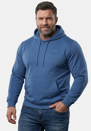Mężczyzna z brodą ubrany w niebieską bluzę z kapturem i dżinsy, uśmiechający się, z jedną ręką w kieszeni na białym tle.