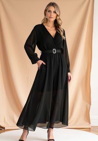 Czarna maxi sukienka z przezroczystą nakładką, długimi rękawami i dekoltem w stylu kopertowym. Posiada zwężoną talię z zapięciem na klamrę. Gładka tekstura.