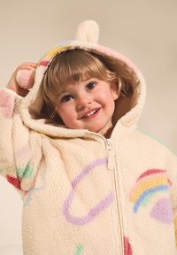 Veste en polaire couleur crème avec motifs arc-en-ciel. Dotée d'une fermeture éclair, d'oreilles d'animal sur la capuche et d'accents en pompons. Texture douce.