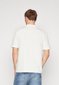 Camisa polo crema con cuello y mangas cortas, hecha de una tela ligera, combinada con pantalones cortos de mezclilla azules.
