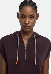 M�łoda kobieta z warkoczem nosząca ciemnofioletową bezrękawnikową bluzę z przednim zamkiem i białymi sznurkami.