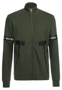 Giacca verde con zip presenta un colletto alto, due tasche frontali con zip e accenti in tessuto nero con testo bianco lungo le maniche.