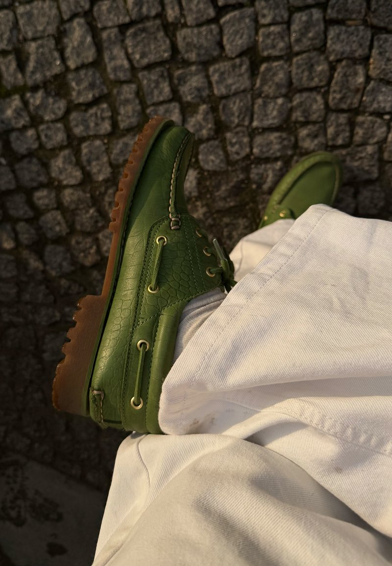 Chaussures bateau en cuir vert avec semelles marron portées avec un pantalon beige clair sur un pavé en pierre.