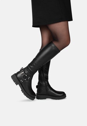 Femme portant des bottes en cuir noires montantes jusqu'aux genoux avec des semelles épaisses et des boucles, associées à des collants noirs transparents et une jupe noire.