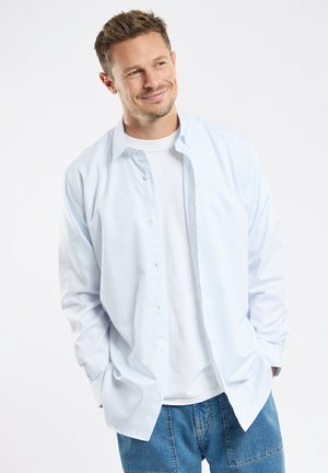 Chemise bleu clair à boutons, manches longues, en tissu léger, portée sur un t-shirt blanc. Coupe décontractée, présentée avec un short en jean.