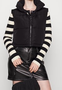 Czarna pikowana kamizelka z wysokim kołnierzem, zapinana na zamek. Pasiasty czarno-kremowy longsleeve, w zestawieniu z czarną skórzaną mini spódnicą.