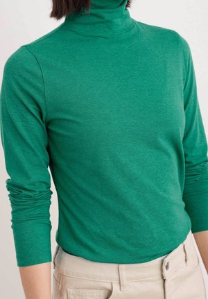 Zielony golf z długim rękawem wykonany z miękkiego materiału, o dopasowanym kroju i lekkim rozciągnięciu. W zestawie z jasnymi spodniami.