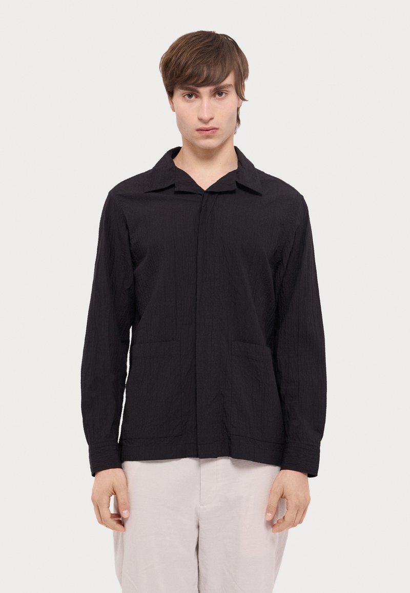 Black long-sleeve shirt made of textured fabric, featuring a collar, front placket, and two side pockets. Minimalist design.