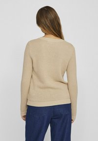 Bež pleteni pulover s teksturiranim uzorkom, okruglim izrezom i rebrastim manžetama i rubovima, složen preko tamnoplave traperice.